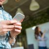Closeup of hands of young man in checkered shirt using mobile phone while his partners arguing