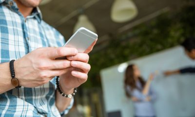 Closeup of hands of young man in checkered shirt using mobile phone while his partners arguing
